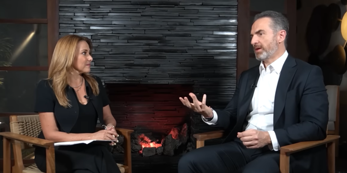 Two professionally dressed people sit in wooden chairs facing each other in a studio by a lit stone fireplace, mid-conversation, microphones visible on their collars.