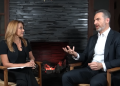 Two professionally dressed people sit in wooden chairs facing each other in a studio by a lit stone fireplace, mid-conversation, microphones visible on their collars.