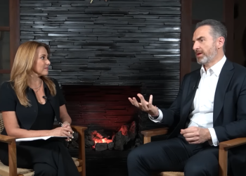 Two professionally dressed people sit in wooden chairs facing each other in a studio by a lit stone fireplace, mid-conversation, microphones visible on their collars.
