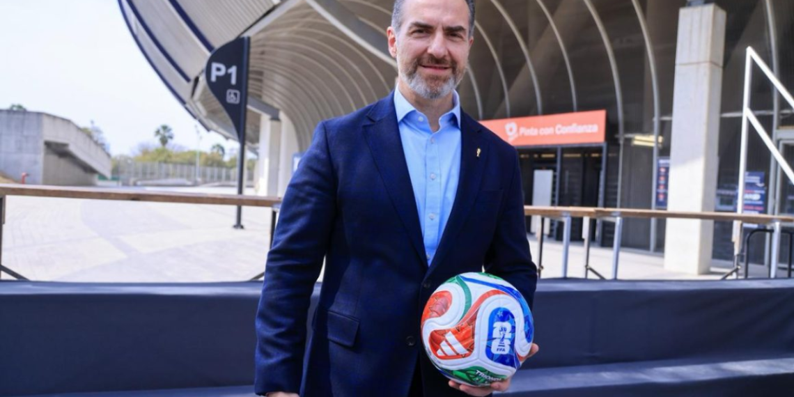 Man in a dark blue suit holding a colorful soccer ball in front of a modern stadium backdrop.