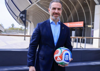 Man in a dark blue suit holding a colorful soccer ball in front of a modern stadium backdrop.