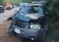 Front of a gray SUV with severe front-end damage after a collision on a rural highway; hood crumpled, windshield damaged, debris on the ground, emergency responders and other vehicles in the background.