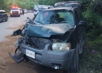 Front of a gray SUV with severe front-end damage after a collision on a rural highway; hood crumpled, windshield damaged, debris on the ground, emergency responders and other vehicles in the background.