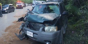 Front of a gray SUV with severe front-end damage after a collision on a rural highway; hood crumpled, windshield damaged, debris on the ground, emergency responders and other vehicles in the background.
