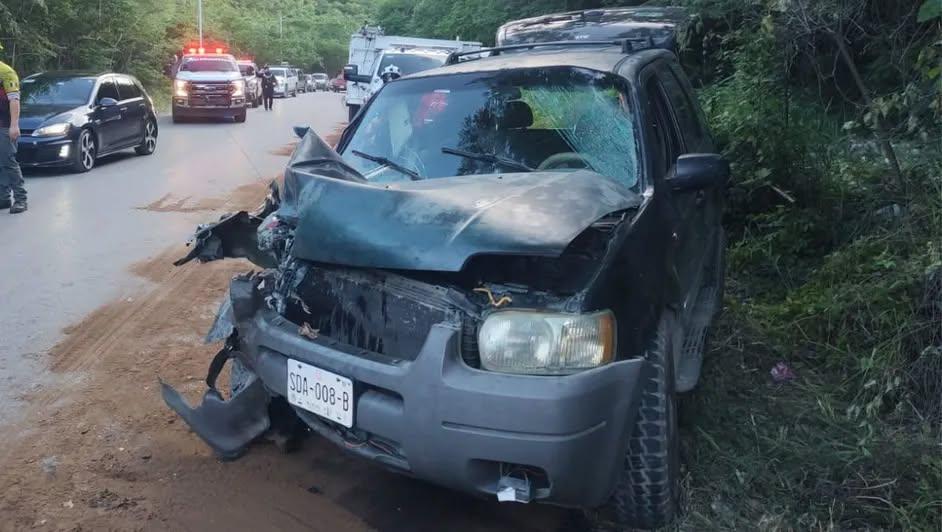 Front of a gray SUV with severe front-end damage after a collision on a rural highway; hood crumpled, windshield damaged, debris on the ground, emergency responders and other vehicles in the background.