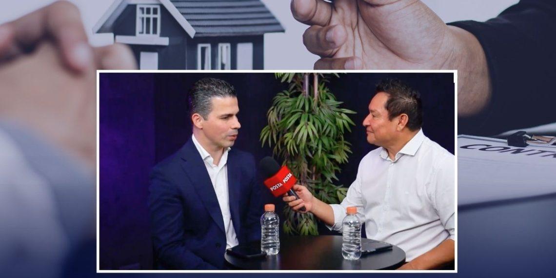 Two men sit at a small round table during an interview; a reporter holds a red POSTA microphone toward the guest, with water bottles on the table and a plant in the background.