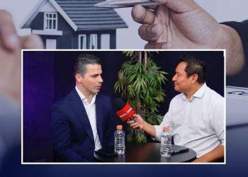 Two men sit at a small round table during an interview; a reporter holds a red POSTA microphone toward the guest, with water bottles on the table and a plant in the background.