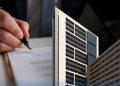 Close-up of a hand signing a document with a miniature modern skyscraper model in the foreground, implying real estate or architecture contracts.
