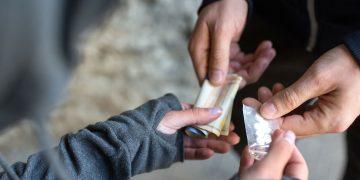 Close-up of hands exchanging cash for a small transparent bag of white pills.