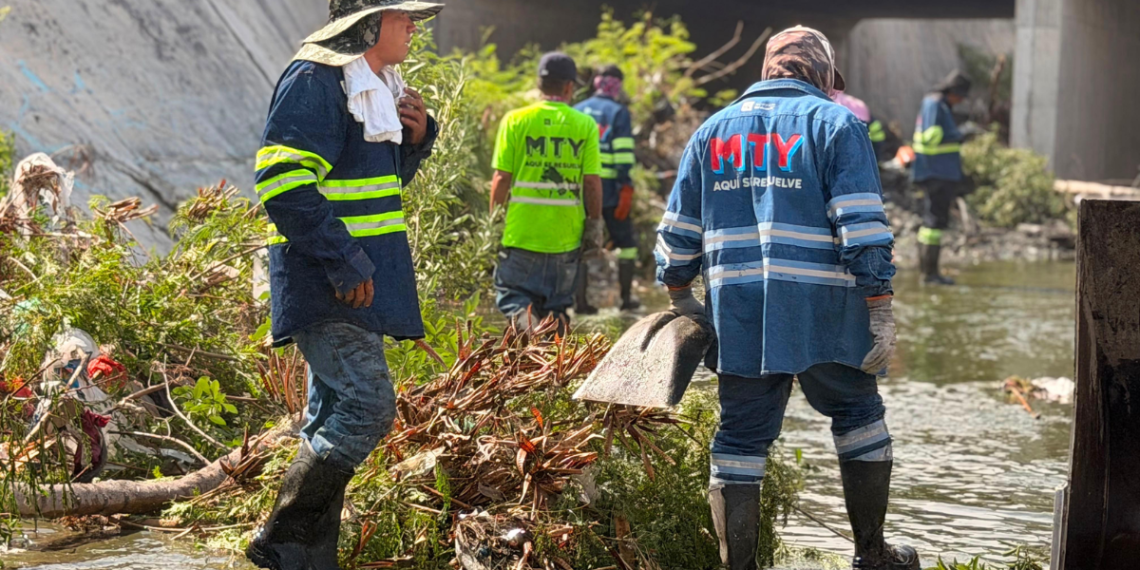 Rescue workers in blue rain gear and reflective stripes stand in floodwater amid debris under a bridge, preparing to clear debris.