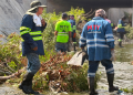 Rescue workers in blue rain gear and reflective stripes stand in floodwater amid debris under a bridge, preparing to clear debris.