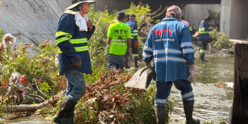 Rescue workers in blue rain gear and reflective stripes stand in floodwater amid debris under a bridge, preparing to clear debris.