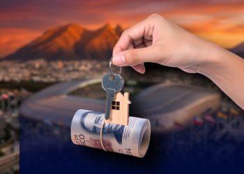 Hand holding a key with a small wooden house charm, suspended over a rolled banknote against a sunset city backdrop (home ownership/mortgage concept).
