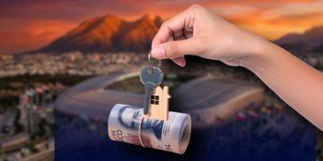 Hand holding a key with a small wooden house charm, suspended over a rolled banknote against a sunset city backdrop (home ownership/mortgage concept).