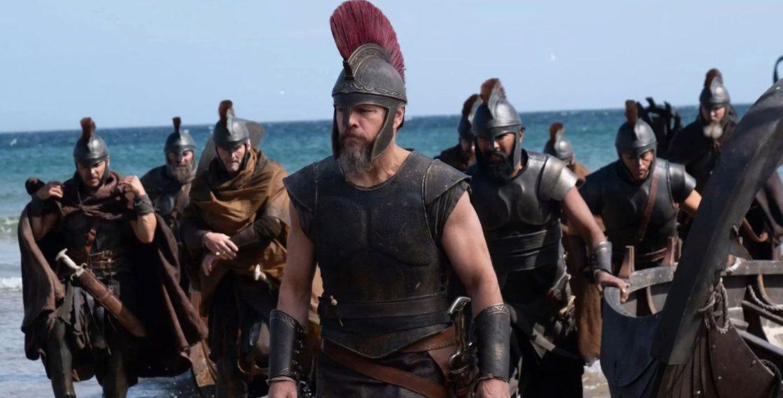 Led by a Spartan-like warrior with a red-crest helmet, armored soldiers march onto a sandy beach with the sea in the background.