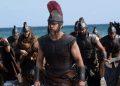 Led by a Spartan-like warrior with a red-crest helmet, armored soldiers march onto a sandy beach with the sea in the background.