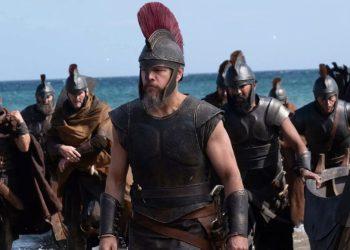 Led by a Spartan-like warrior with a red-crest helmet, armored soldiers march onto a sandy beach with the sea in the background.