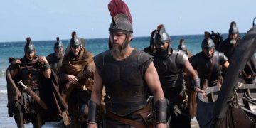 Led by a Spartan-like warrior with a red-crest helmet, armored soldiers march onto a sandy beach with the sea in the background.