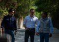 Three men walk along a sunlit, tree‑lined path, chatting as they stride side by side. Center man wears a white shirt and jeans, flanked by others in dark shirts/vests.