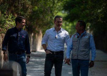 Three men walk along a sunlit, tree‑lined path, chatting as they stride side by side. Center man wears a white shirt and jeans, flanked by others in dark shirts/vests.