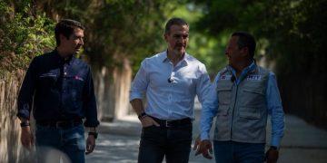 Three men walk along a sunlit, tree‑lined path, chatting as they stride side by side. Center man wears a white shirt and jeans, flanked by others in dark shirts/vests.