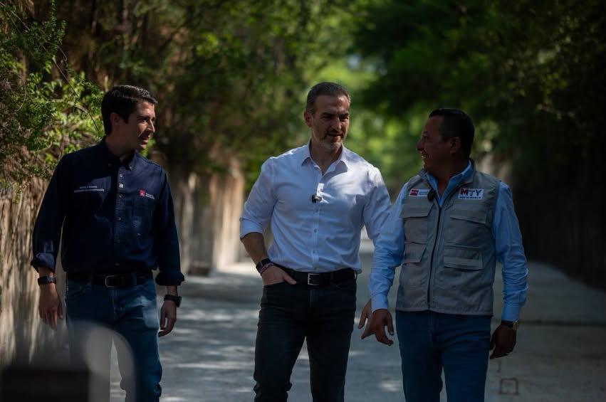 Three men walk along a sunlit, tree‑lined path, chatting as they stride side by side. Center man wears a white shirt and jeans, flanked by others in dark shirts/vests.
