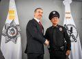 Two men shake hands in front of security agency banners during a formal ceremony—the suited official and a police officer in uniform smile for the photo.