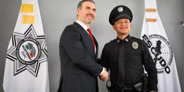 Two men shake hands in front of security agency banners during a formal ceremony—the suited official and a police officer in uniform smile for the photo.