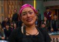 Close-up of a smiling woman wearing a pink headscarf and black apron, in a studio with a seated audience in the background.