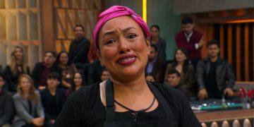 Close-up of a smiling woman wearing a pink headscarf and black apron, in a studio with a seated audience in the background.