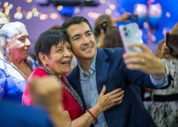 Man in a blue suit takes a smiling selfie with an elderly woman in red at a festive event.