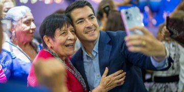 Man in a blue suit takes a smiling selfie with an elderly woman in red at a festive event.