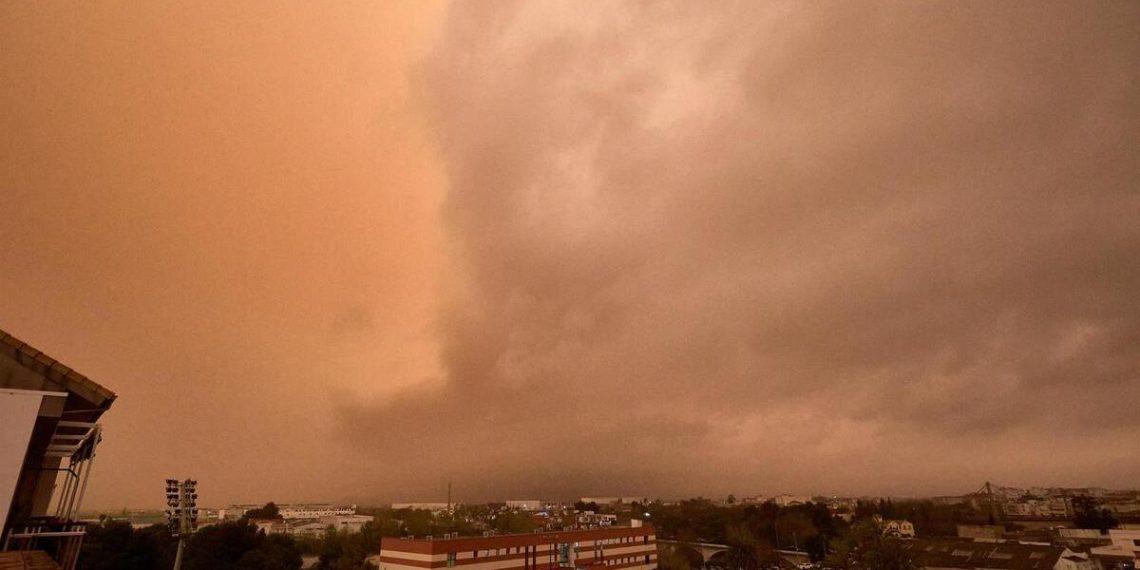 Orange-tinted sky with large cloud front over a city, cars parked in a lot and a brick building in the foreground.