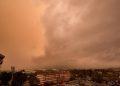 Orange-tinted sky with large cloud front over a city, cars parked in a lot and a brick building in the foreground.