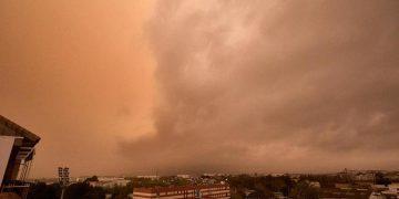 Orange-tinted sky with large cloud front over a city, cars parked in a lot and a brick building in the foreground.